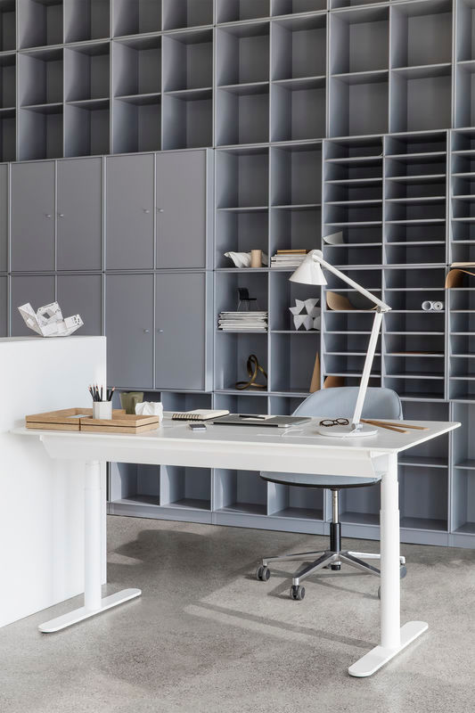 Weißer Hilow Tisch, höhenverstellbar, mit Schreibtischlampe, Laptop und Büromaterial vor grauem Regalsystem.