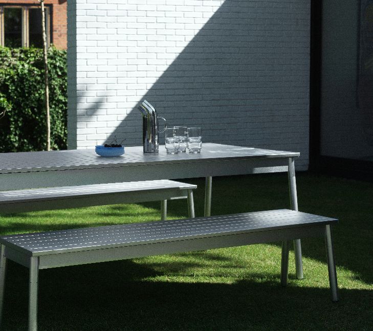 Normann Copenhagen Ex Bank Outdoor auf Rasen mit Tisch, Bank, Getränken und Backsteinhintergrund.