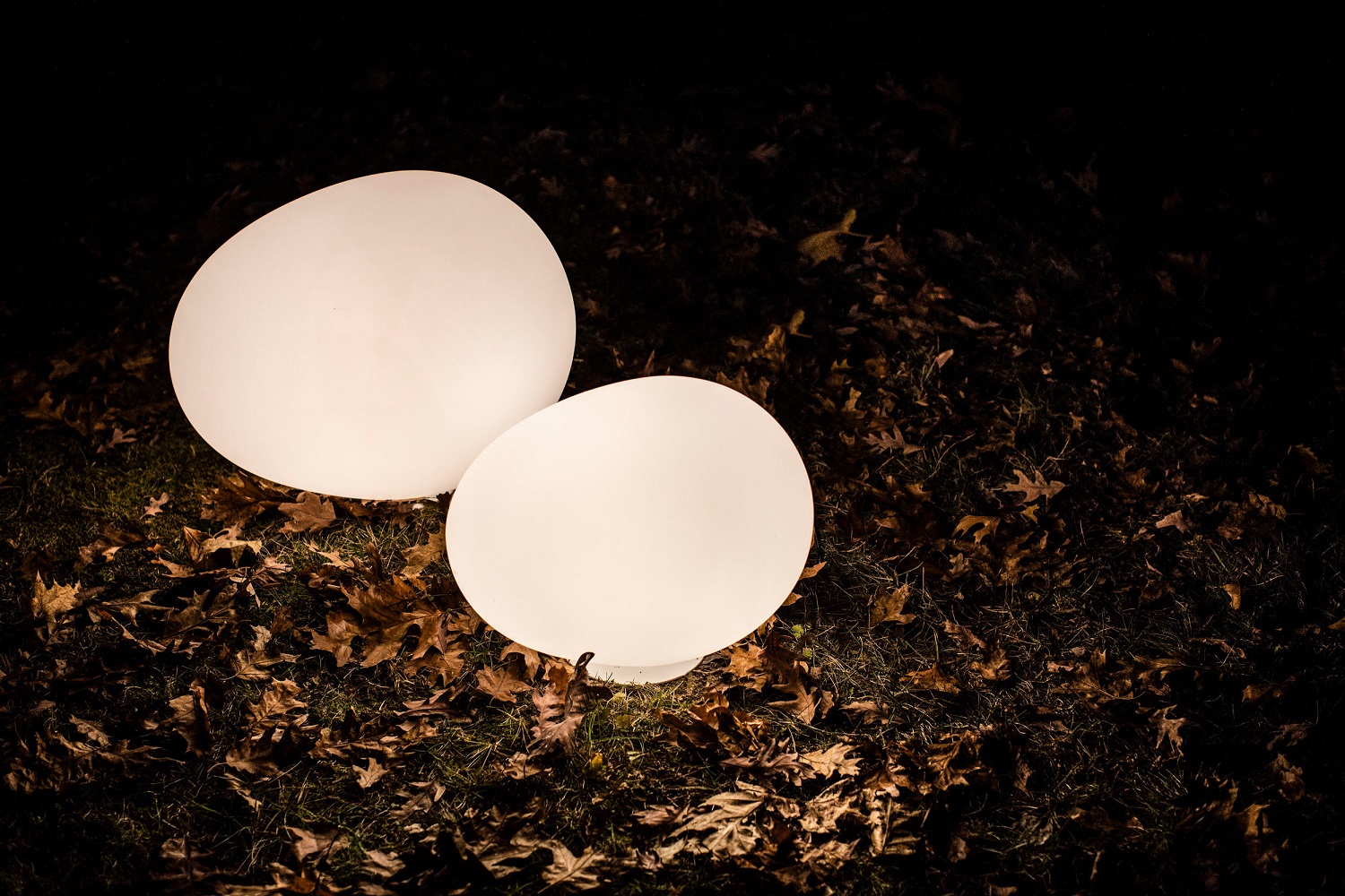 Zwei leuchtende Gregg Outdoor Bodenleuchten von Foscarini auf Rasen mit Herbstlaub im Dunkeln.