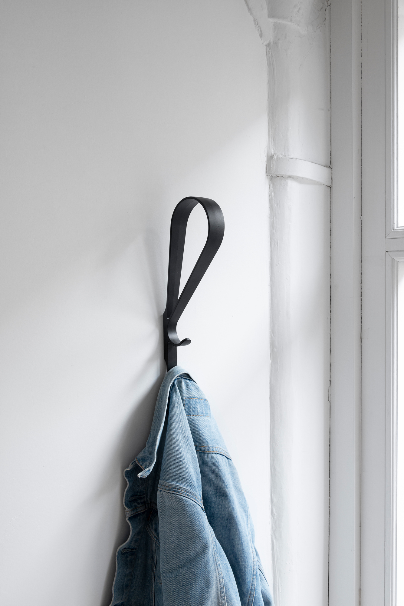 Schwarzer Artek Tupla Wandhaken an weißer Wand mit hängender Jeansjacke, moderne Garderobenlösung für Flur und Zuhause.