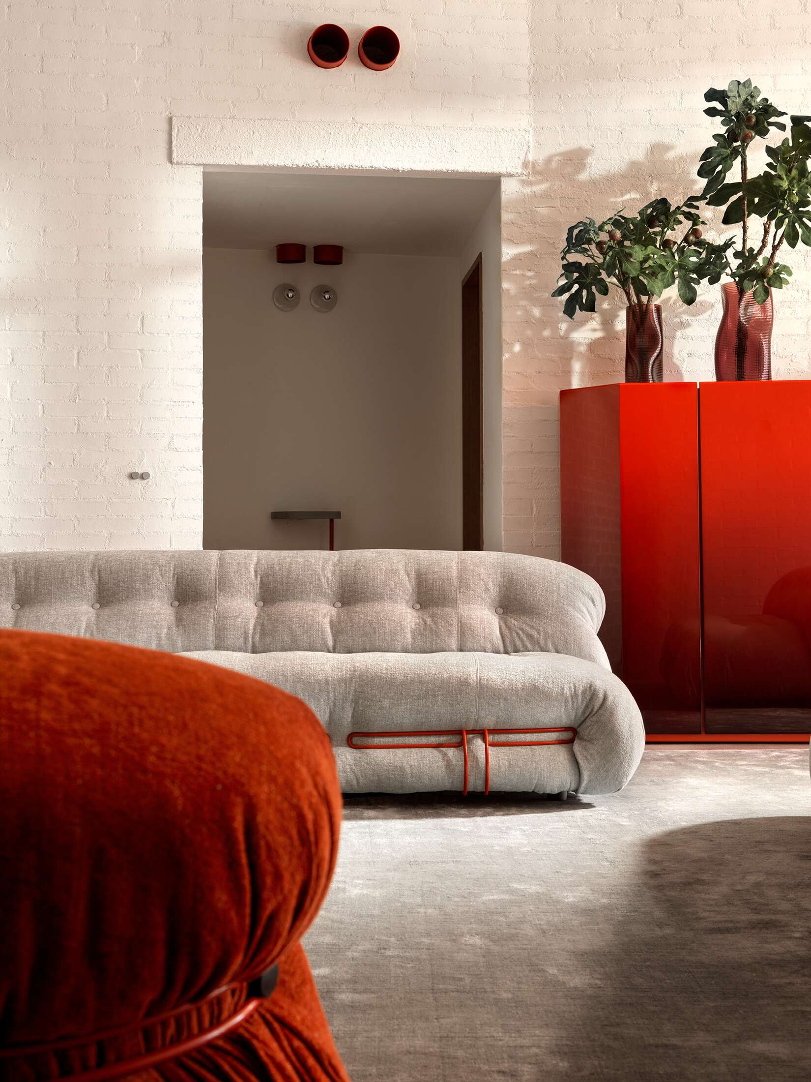 Helles Wohnzimmer mit grauem Cassina Sofa, rotem Sessel und Schrank, Pflanzen und weißer Ziegelwand.