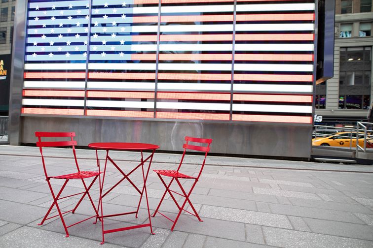 Roter Fermob Bistro Tisch mit zwei Stühlen im urbanen Außenbereich vor US-amerikanischer Flagge.