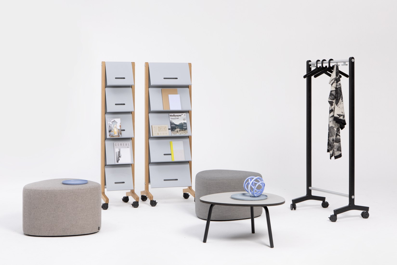 Zwei Round20 Prospekthalter von Cascando mit Zeitschriften, Hocker, Tisch und Kleiderständer auf weißem Hintergrund.