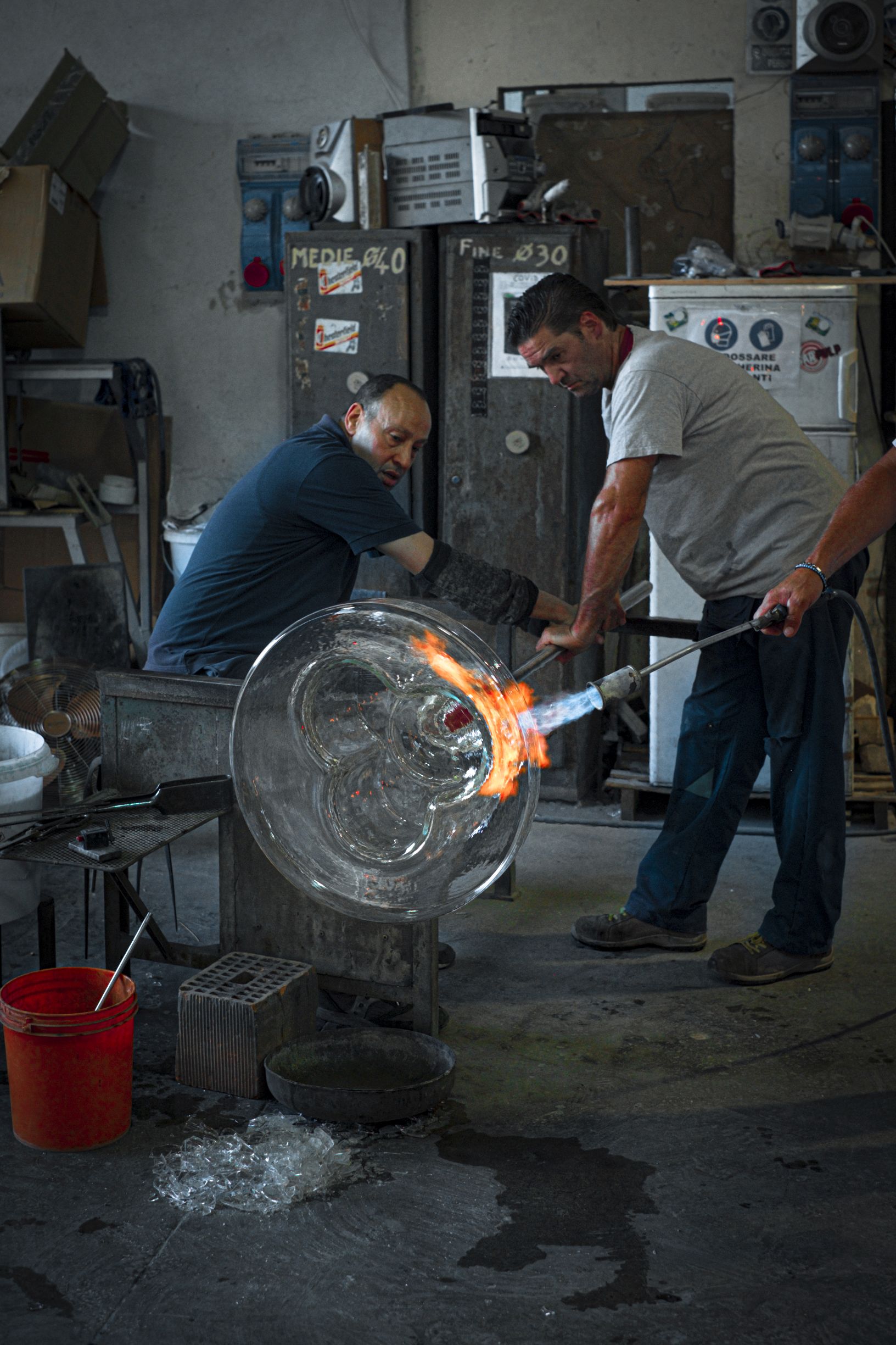 Zwei Glasbläser formen eine große Glasschale mit Feuer in einer Werkstatt.