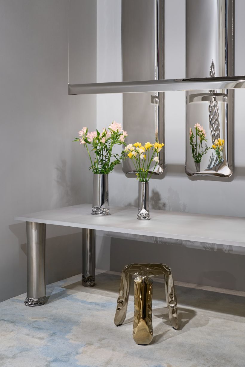 Silberne Designervasen mit Blumen auf weißem Tisch, dazu passender Hocker und Wanddekoration.