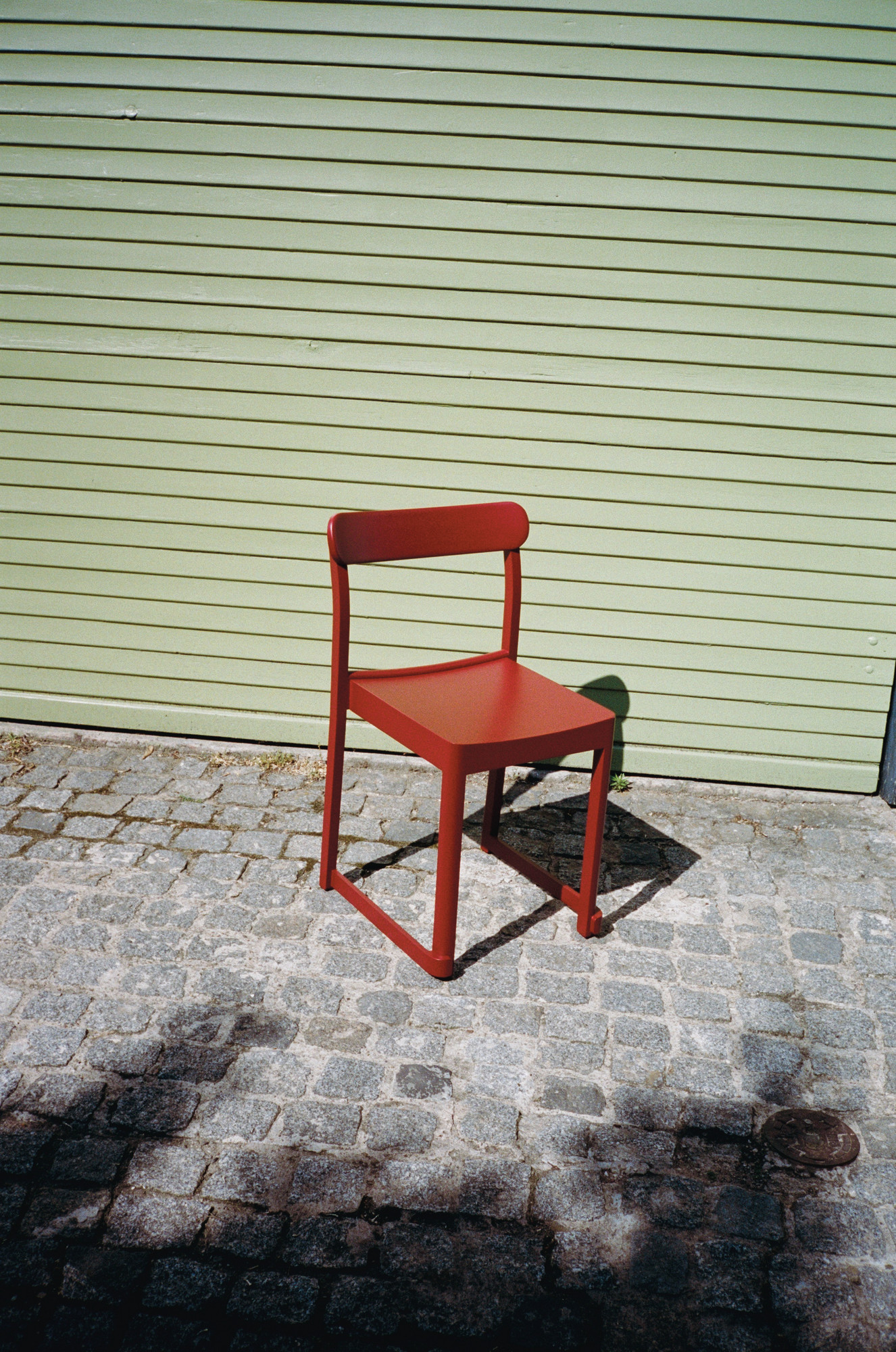 Roter Atelier Chair Stuhl von Artek vor einer grünen Holzwand auf Pflastersteinen.