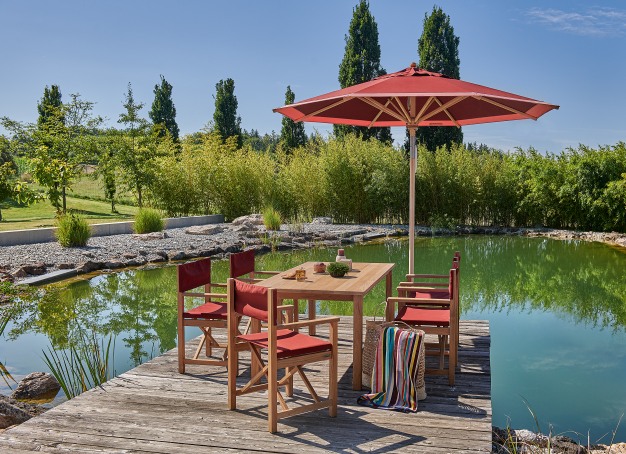 Weishäupl Klassiker Sonnenschirm und Gartenmöbel Set aus Holz mit rotem Schirm und Stühlen am See.