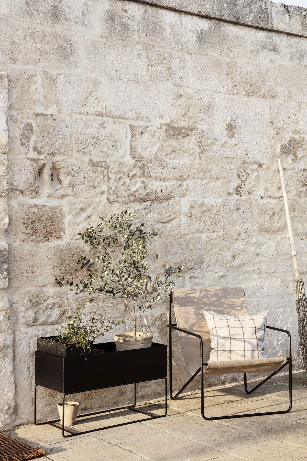 Ferm Living Desert Chair Loungesessel mit Kissen und schwarzem Pflanzenkasten vor einer Steinwand.