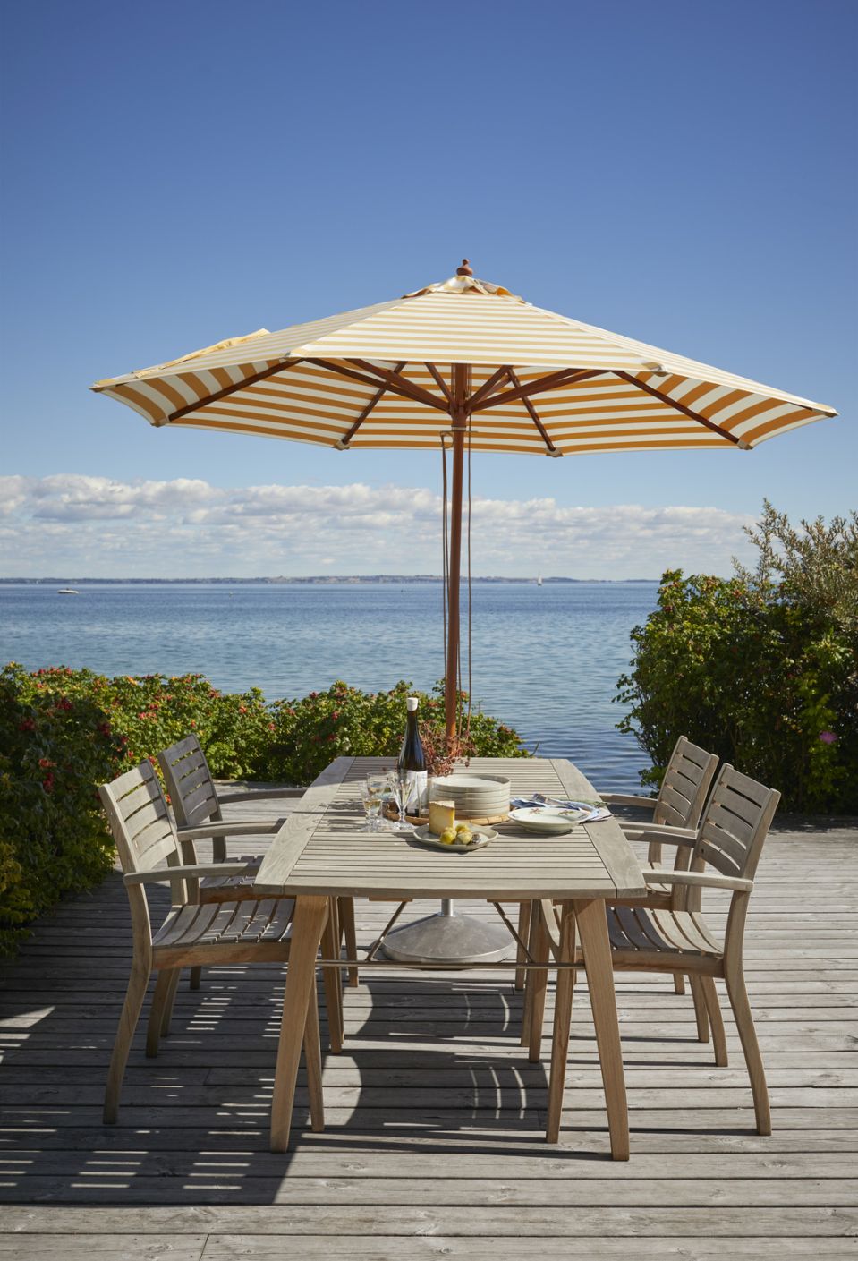 Ballare Chair von Skagerak by Fritz Hansen, Outdoor-Szene mit Tisch, Stühlen und Sonnenschirm am Meer.