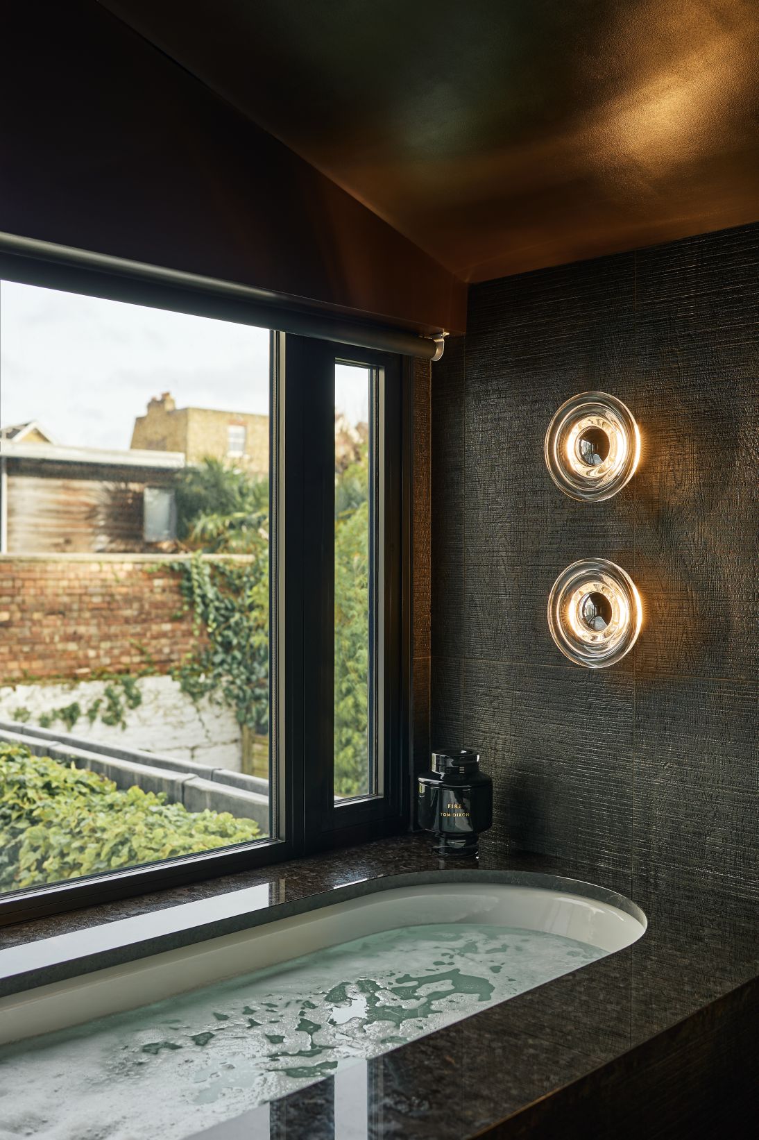 Zwei Tom Dixon Press Surface Wandleuchten neben einem Fenster über einer Badewanne im Badezimmer.