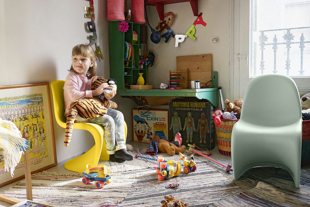 Mädchen sitzt auf gelbem Kinderstuhl im Kinderzimmer mit Spielzeug und farbenfroher Einrichtung. Design Kinderstuhl.