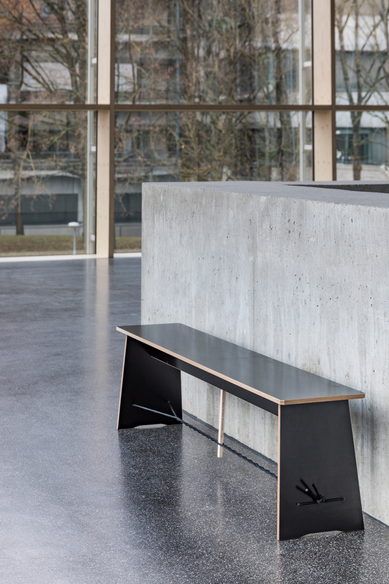 Lange, schwarze Sitzbank "Langer Max" von Nils Holger Moormann vor einer Betonwand und Fensterfront.