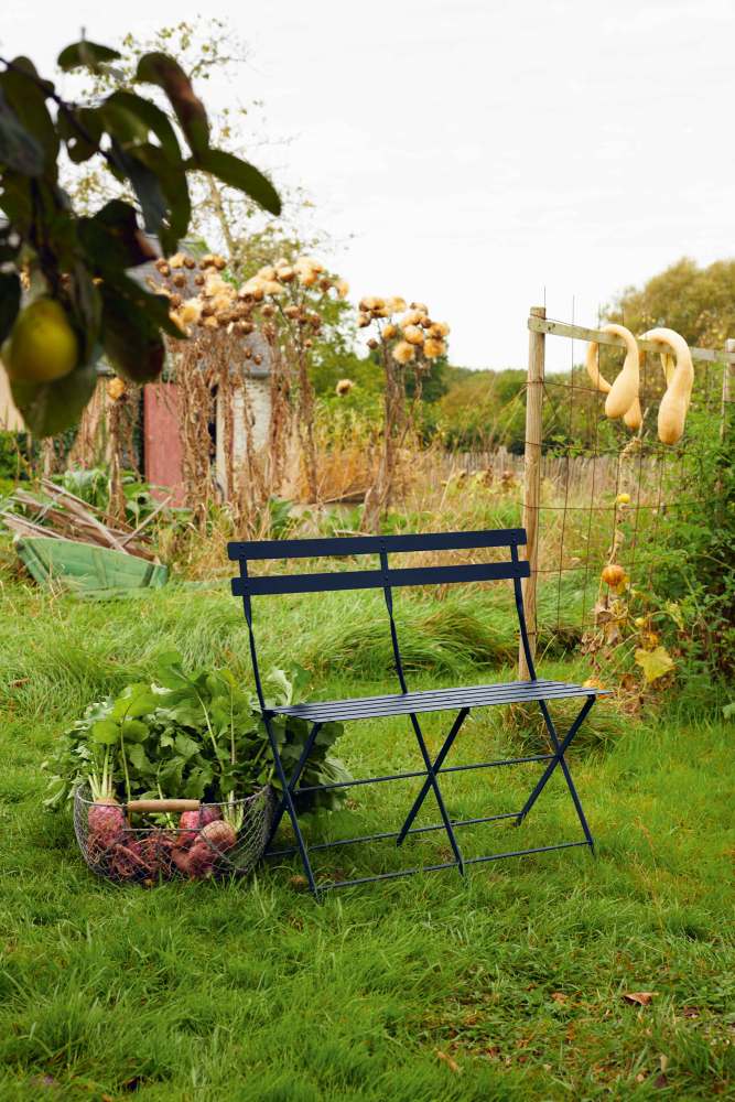 Blaue Fermob Bistro Klappbank für zwei Personen im Garten, neben einem Korb mit Gemüse.