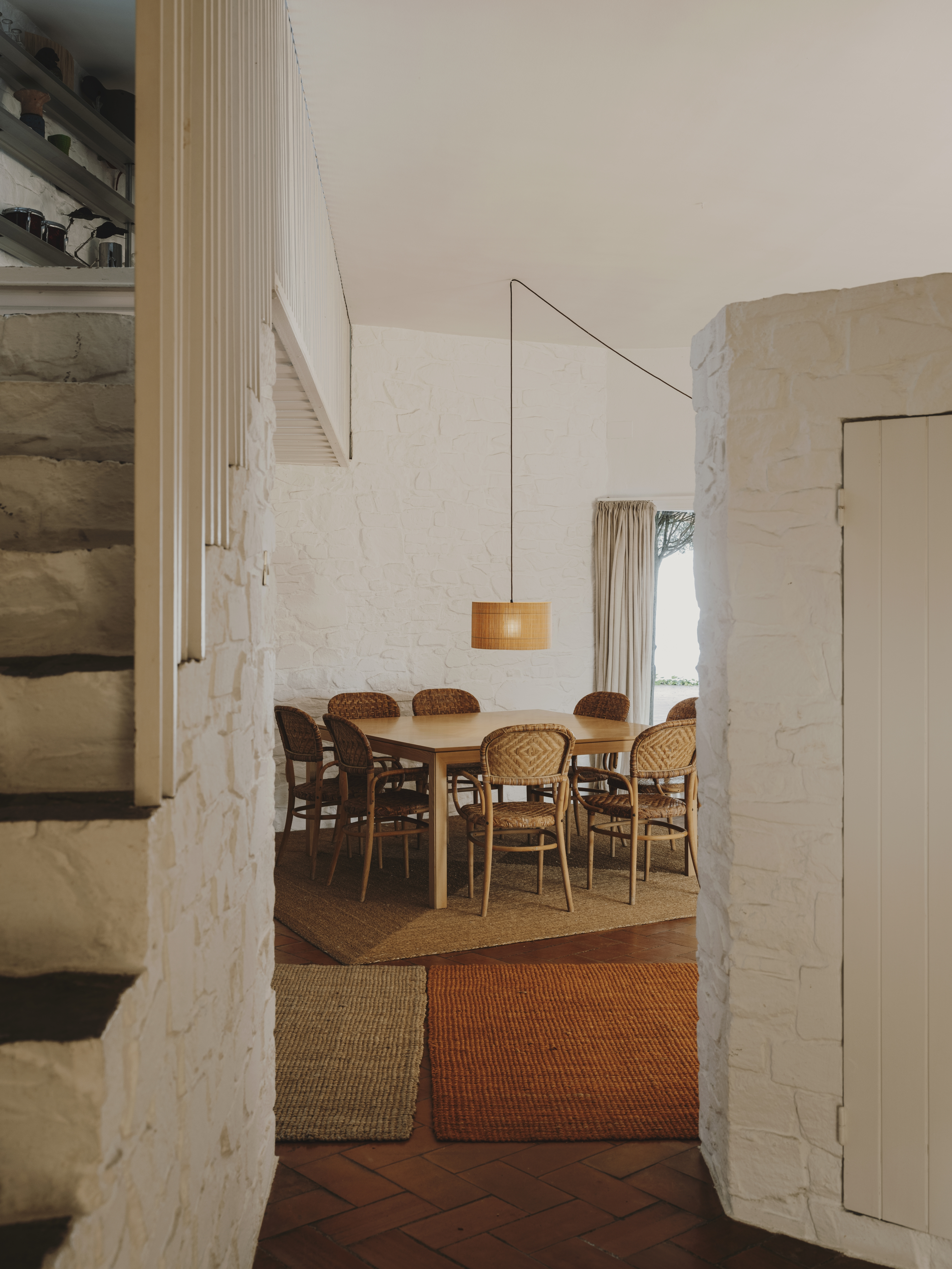 Esszimmer mit Holztisch, Rattan-Stühlen und gewebter Pendelleuchte in einem Haus mit weißen Ziegelwänden.