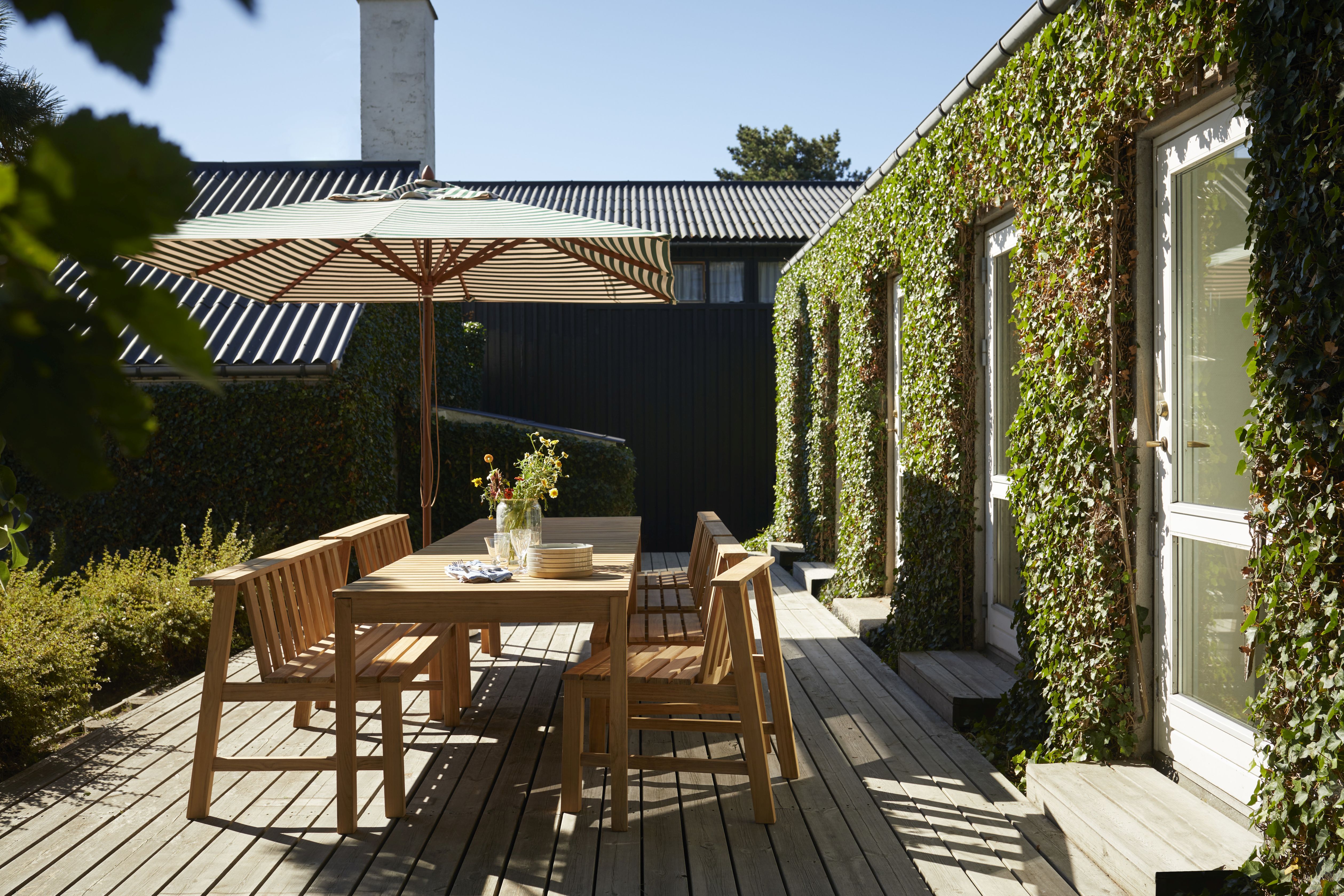 Cadence Bank Teak Gartengarnitur von Skagerak by Fritz Hansen auf Holzterrasse mit Sonnenschirm und Bewuchs.