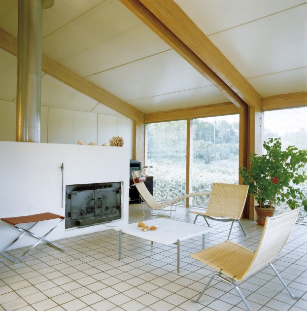 Heller Wohnraum mit PK 61 Couchtisch von Fritz Hansen, Stühlen, Liege und großem Fenster mit Blick ins Grüne.