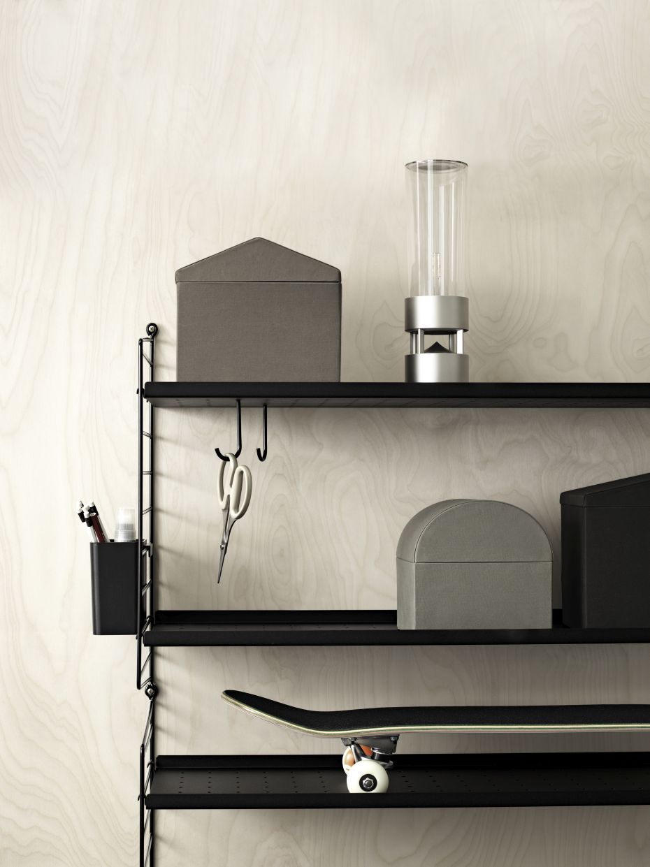 Schwarzes Metallregal mit String Furniture Haken, Boxen, Lampe und Skateboard vor hellem Hintergrund.