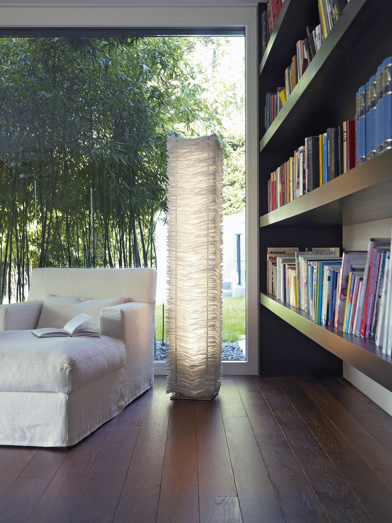 Moderne Stehlampe aus Papier in Wohnzimmer mit Sofa und Bücherregal, Tageslicht.