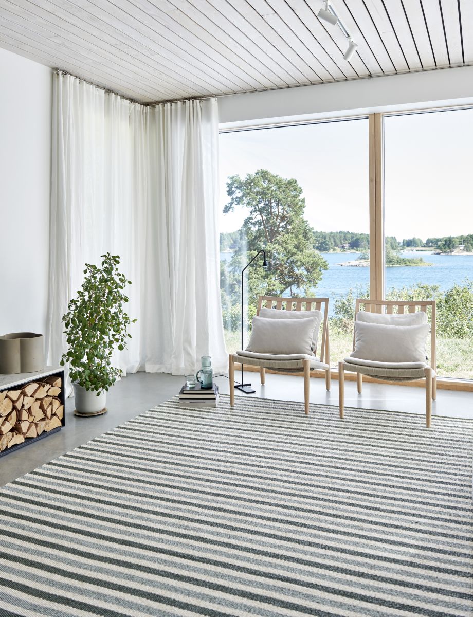 Teo Teppich 230x320cm Pappelina im hellen Wohnzimmer mit Streifenmuster, Stühlen und großem Fenster.