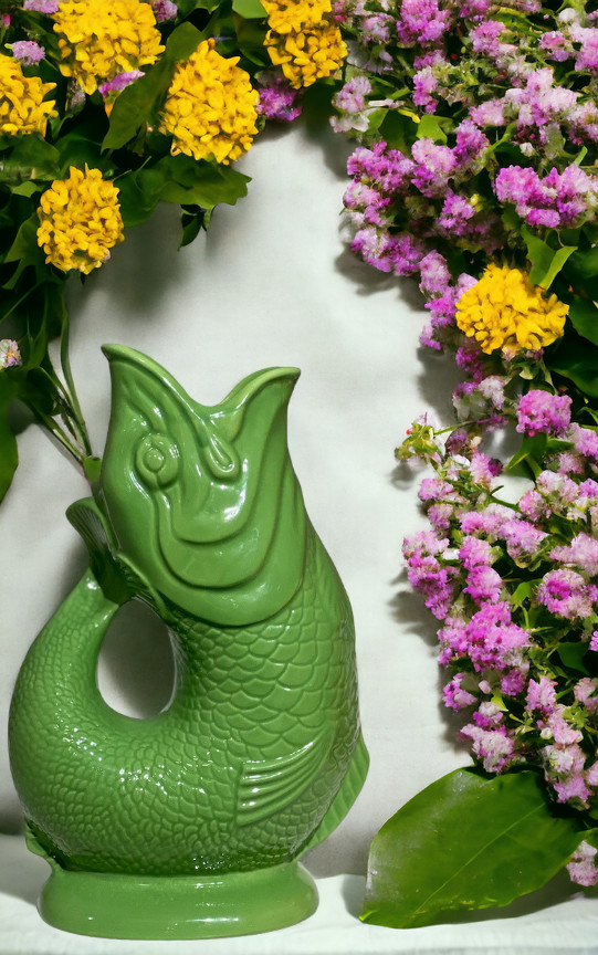 Grüne Gluckigluck Fischkaraffe XL als Vase, umgeben von gelben und lila Blumen.