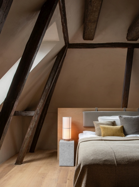 Audo Hashira Tischleuchte auf Nachttisch im Schlafzimmer mit Holzbalken und Bett.