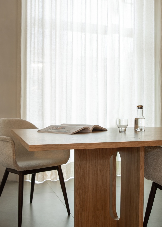 Androgyne Dining Table Esstisch aus Eiche mit Stühlen, Wasserflasche und Magazin vor hellem Vorhang.