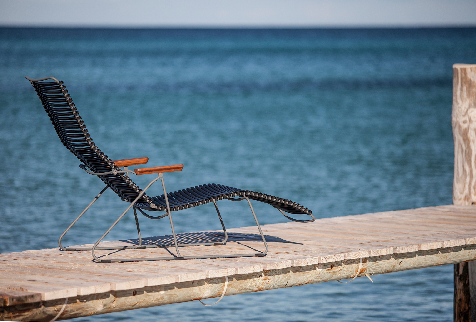 Click Sunlounger Outdoor Sonnenliege von Houe in Dunkelgrau auf einem Holzsteg mit Meerblick.