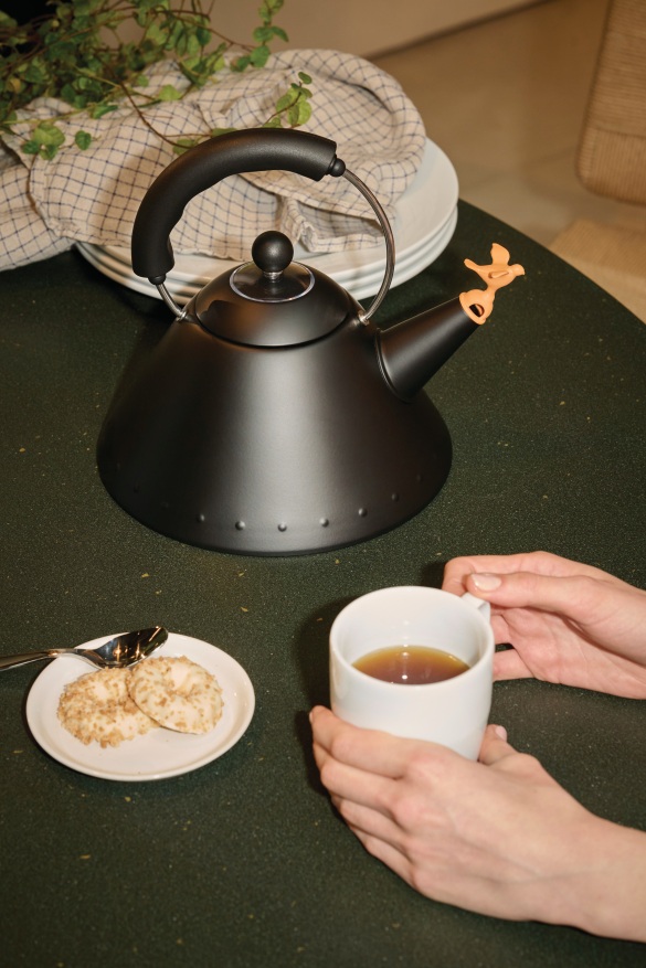 Schwarzer Alessi 9093 BB Wasserkessel mit Vogelpfeife, Tasse Tee und Gebäck auf einem Tisch.
