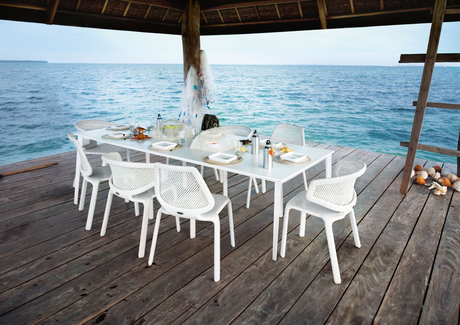 Esstisch mit weißen Design-Stühlen auf Holzsteg am Meer, gedeckt mit Geschirr und Getränken.