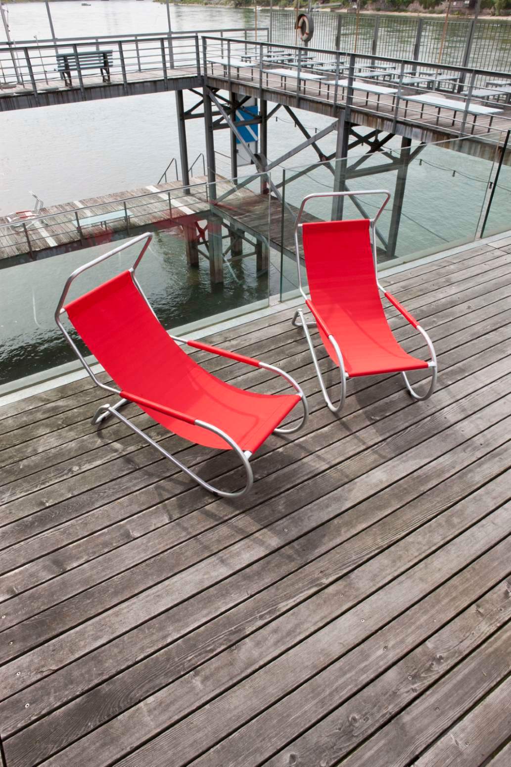 Zwei rote Lido Liegen mit silbernem Gestell auf einer Holzterrasse am Wasser.