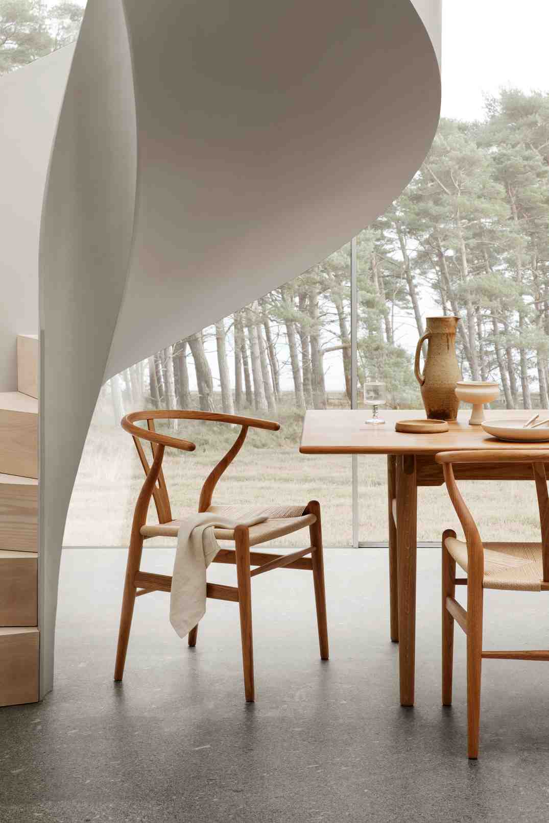 Helle Esszimmer-Szene mit Holz-Esstisch und Wishbone Stühlen mit geflochtenen Sitzen vor großem Fenster.