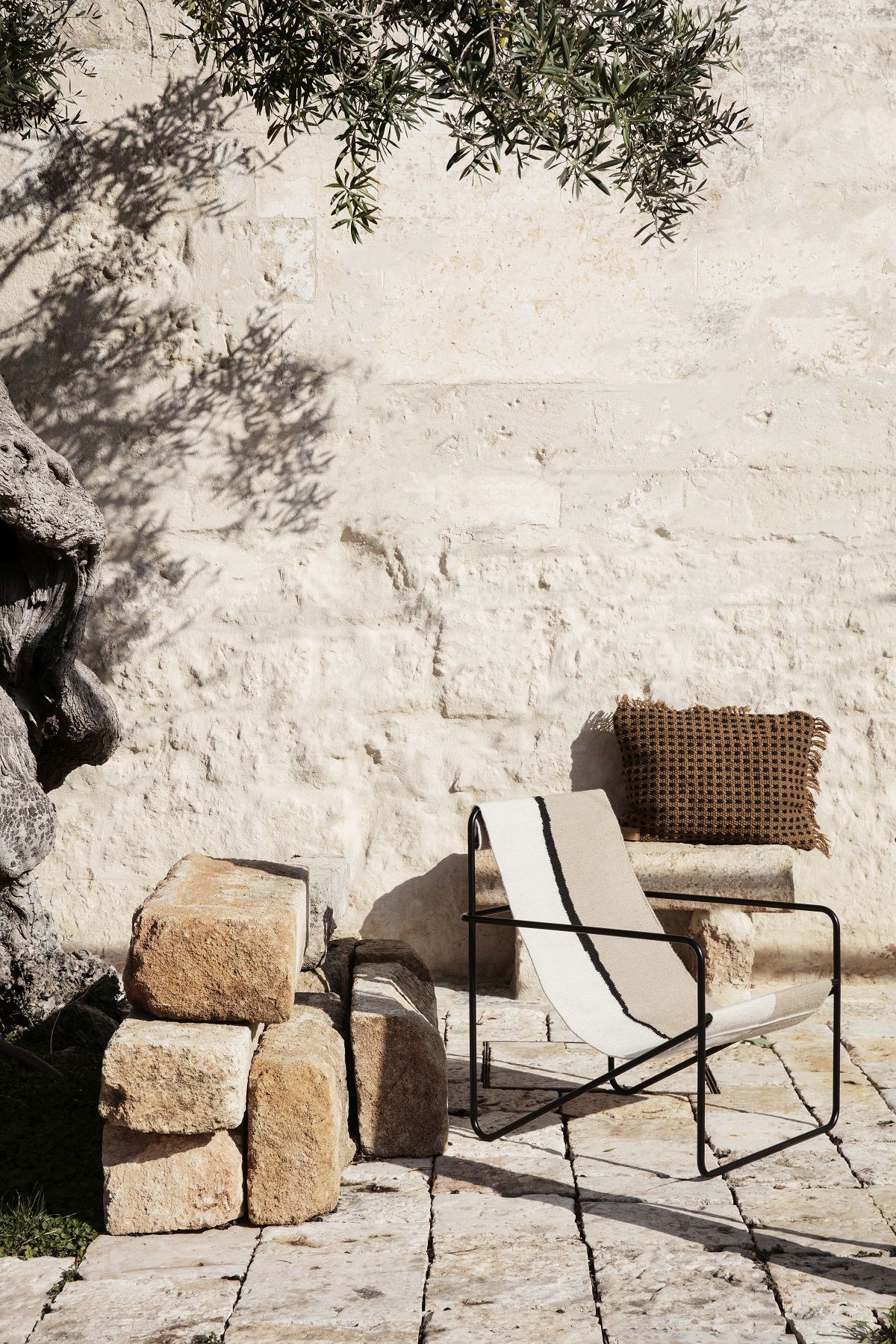 Ferm Living Desert Chair Loungesessel mit Kissen vor einer Steinwand im Außenbereich.