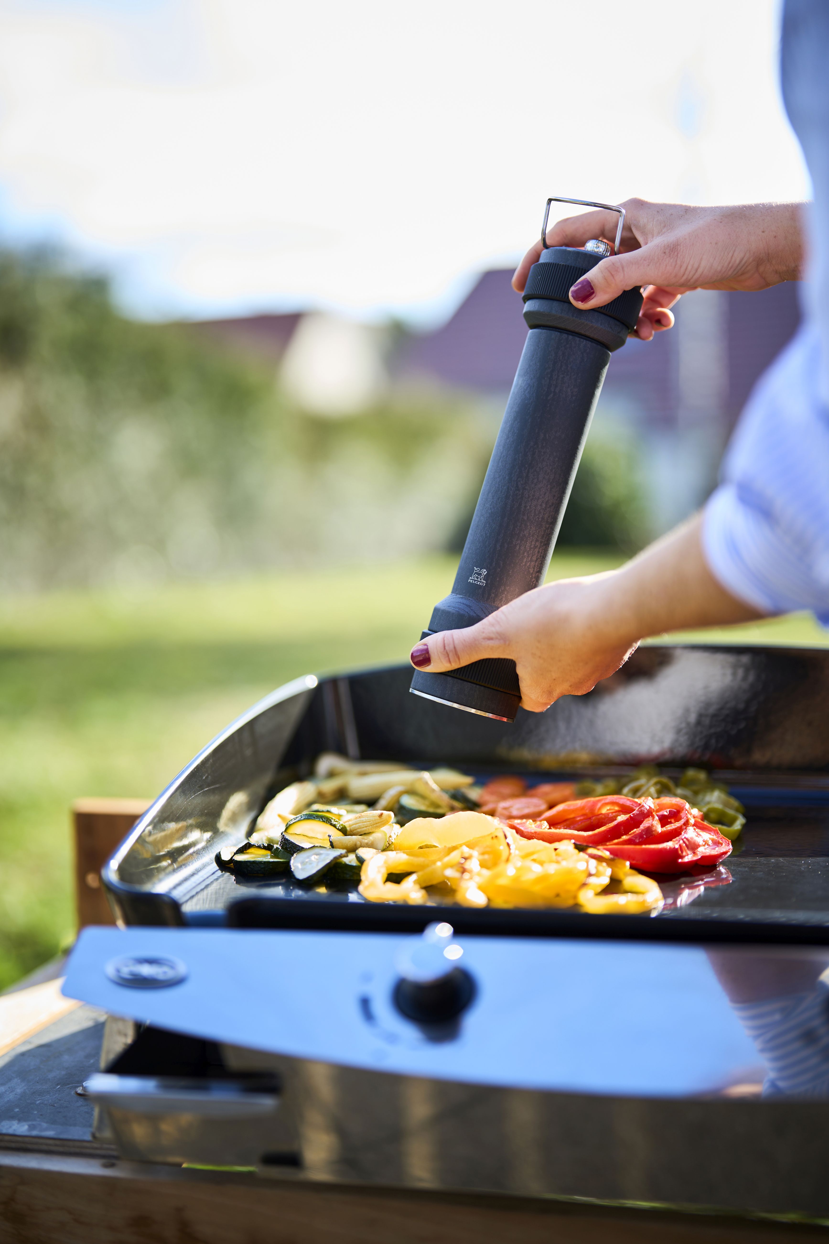 Frau würzt Gemüse auf Grill mit Peugeot BBQ Pfeffermühle Graphit 30 cm im Freien.