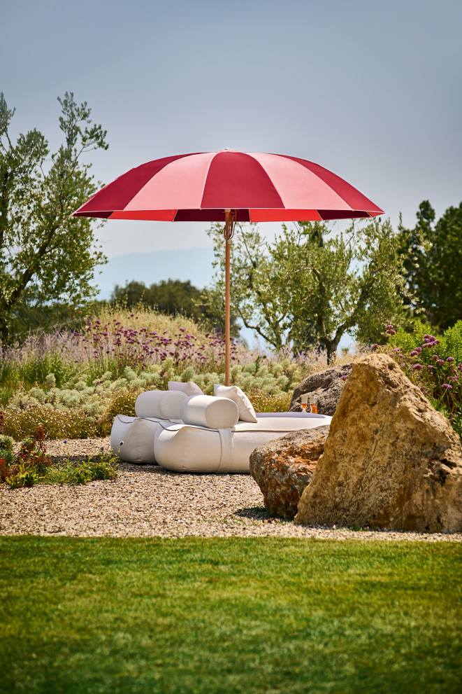Weishäupl Pagodenschirm: Rot-gestreifter Sonnenschirm spendet Schatten über einer weißen Outdoor-Lounge in einem üppigen Garten.