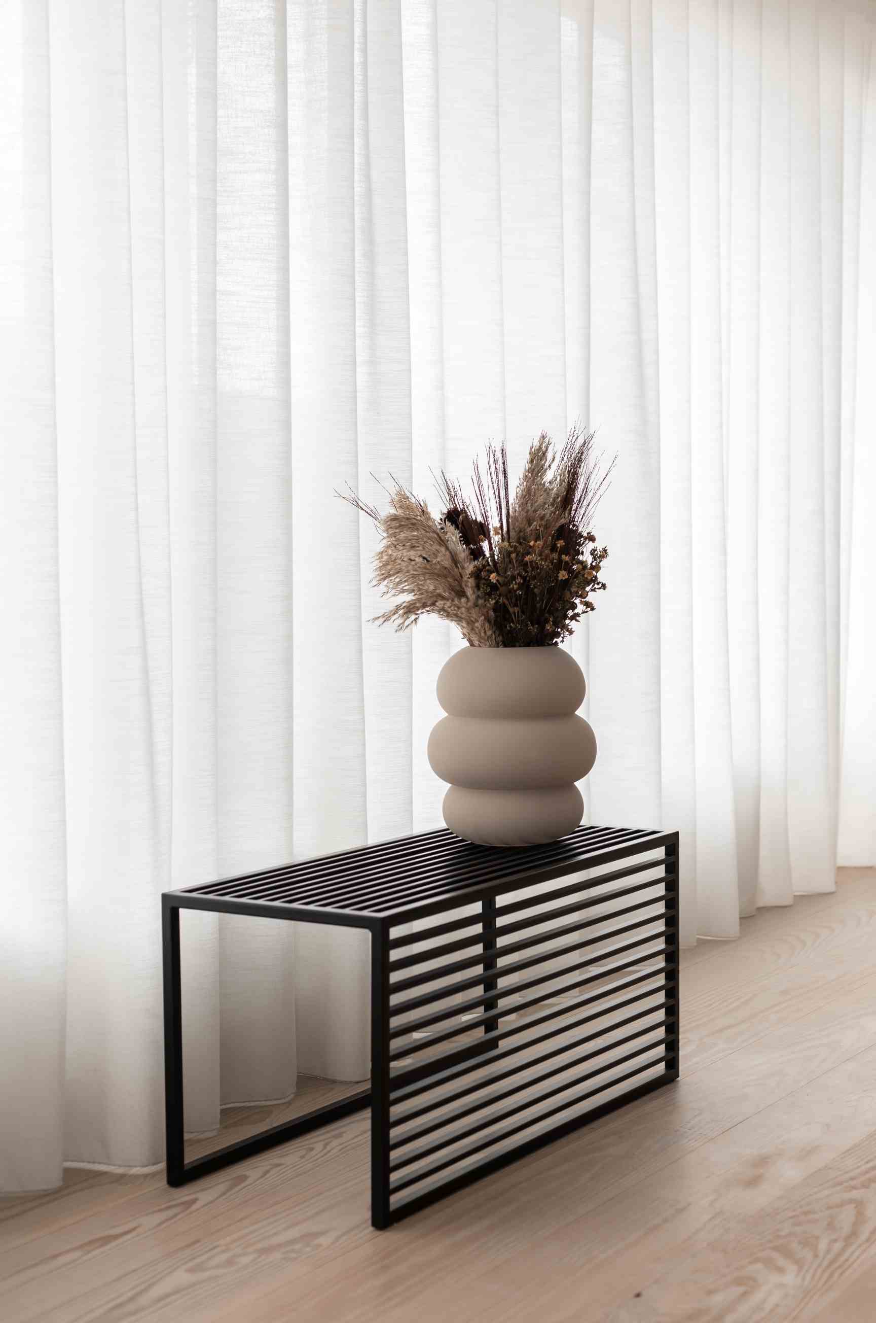 Novoform Element Bank mit schwarzem Metallgestell und beiger Blumenvase mit Trockenblumen vor weißem Vorhang.
