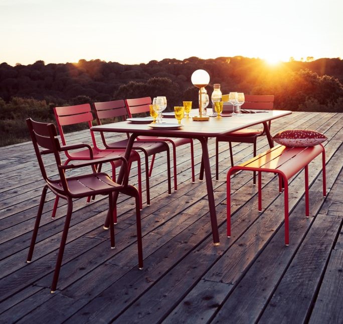Roter Gartentisch mit Stühlen und Bank auf Holzterrasse bei Sonnenuntergang, modernes Gartenmöbel-Set für den Außenbereich.
