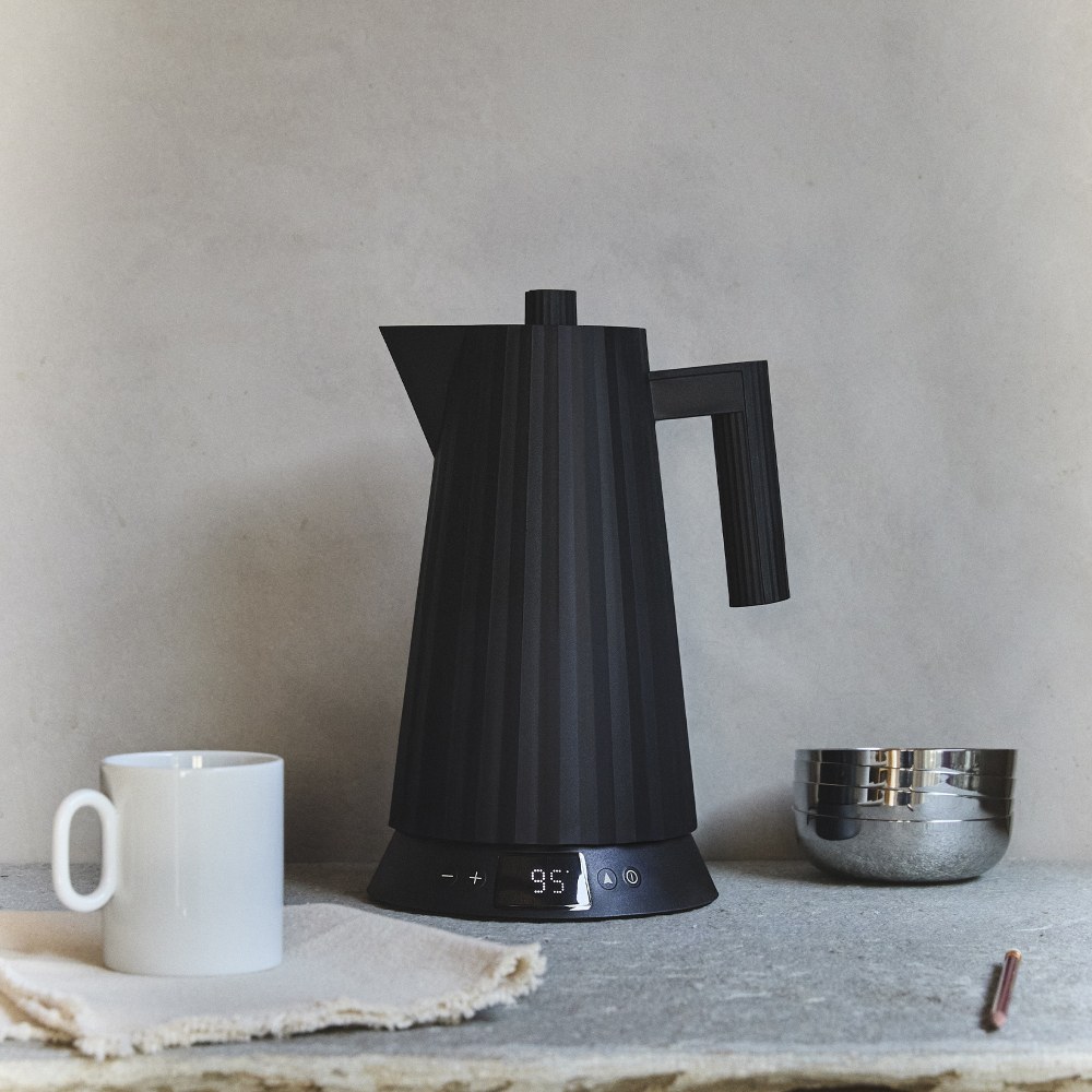 Schwarzer Alessi Plissé Wasserkocher MDL21 mit digitaler Temperaturanzeige neben Tasse und Schale.
