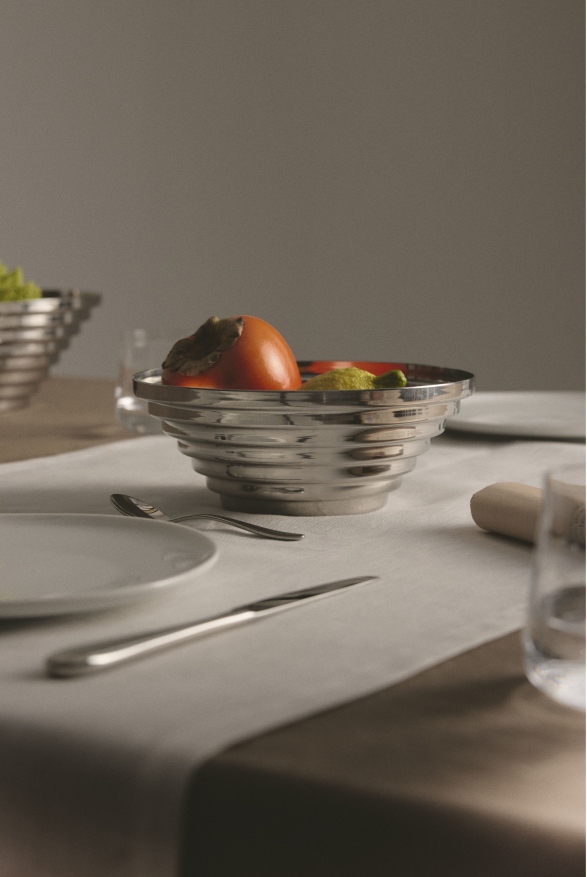 Alessi Maya Schale, runde Designschale aus Edelstahl mit Obst auf gedecktem Tisch.
