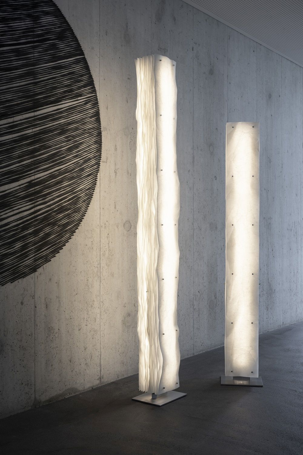 Zwei moderne Totem Stehleuchten aus hellem Stoff und Metallfuß vor Betonwand.