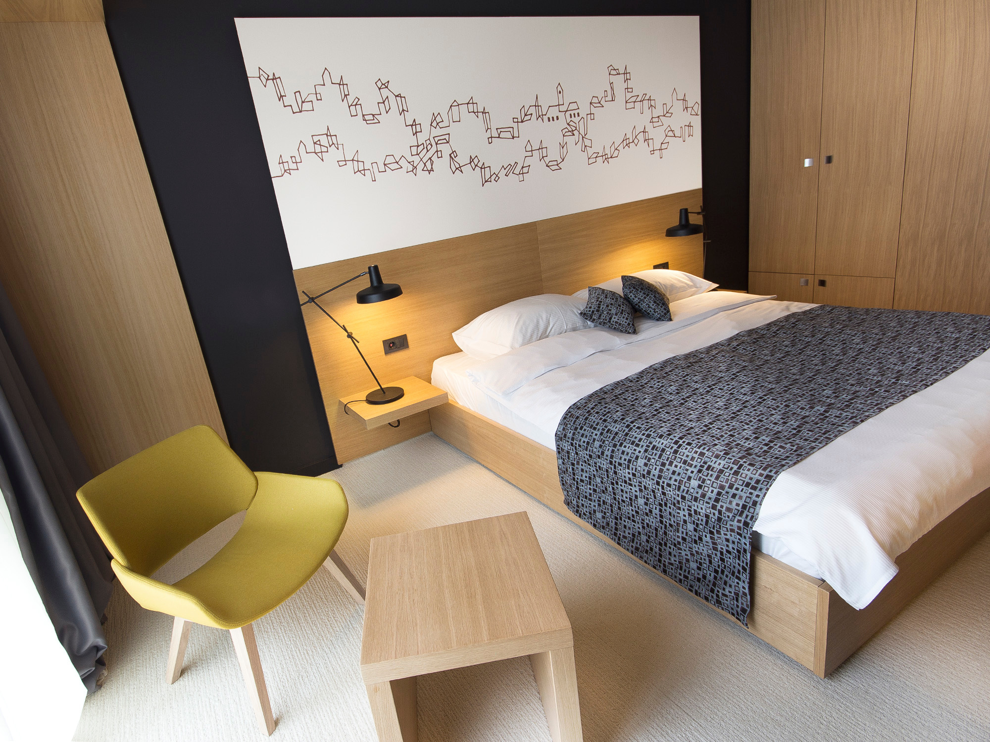 Schwarze Arigato Tischleuchte von Grupa EINZELSTÜCK auf Nachttisch in modernem Hotelzimmer mit Bett und gelbem Sessel.