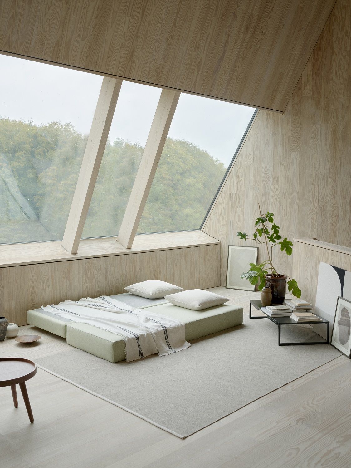 Helles Zimmer mit Lubi Daybed Schlafsofa, zwei Kissen und Decke vor großem Fenster.