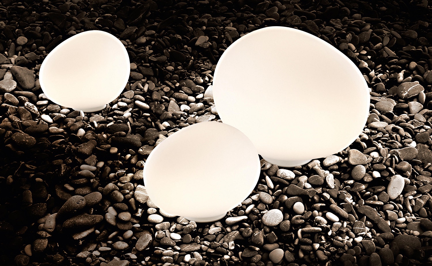 Foscarini Gregg Outdoor Leuchten: Drei eiförmige Bodenleuchten in Weiß auf dunklem Kiesstrand, stimmungsvolle Gartenbeleuchtung.