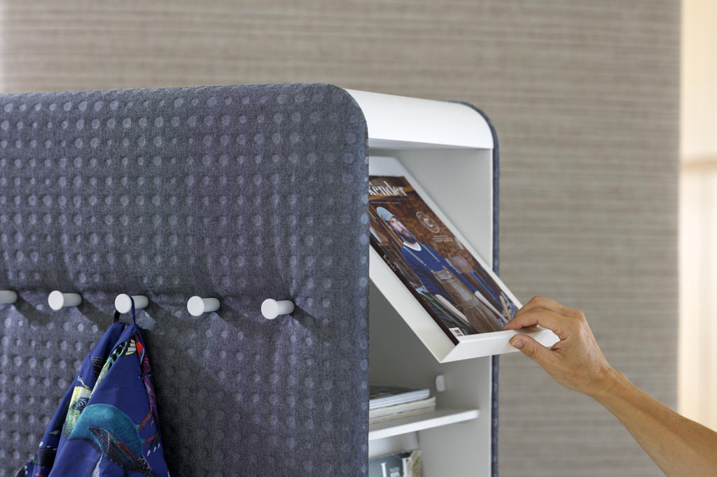 Cascando Pillow Space Akustik-Raumteiler mit Garderobenhaken und Zeitschriftenregal, Hand legt Zeitschrift hinein.