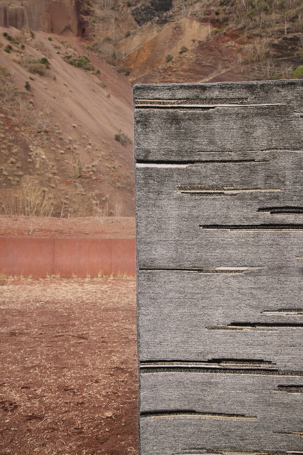 Colorado Teppich von Nanimarquina, grauer Wollteppich mit horizontalen Linien vor roter Landschaft.