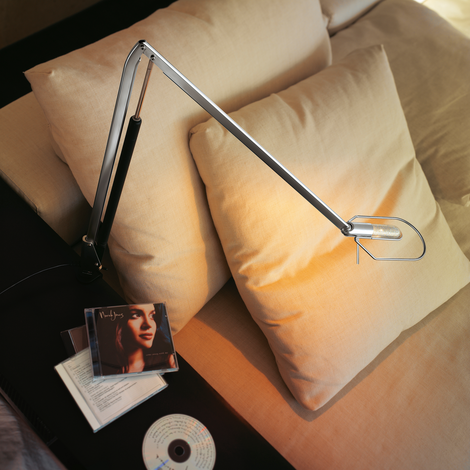 Lifto LED Tischleuchte von Belux, silberfarben, dimmbar, auf einem Sofa mit Kissen platziert.