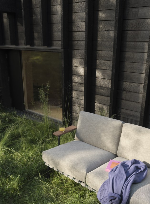 Muuto Settle Outdoor Sofa 2-Sitzer in Beige mit Getränk und Buch im Garten.