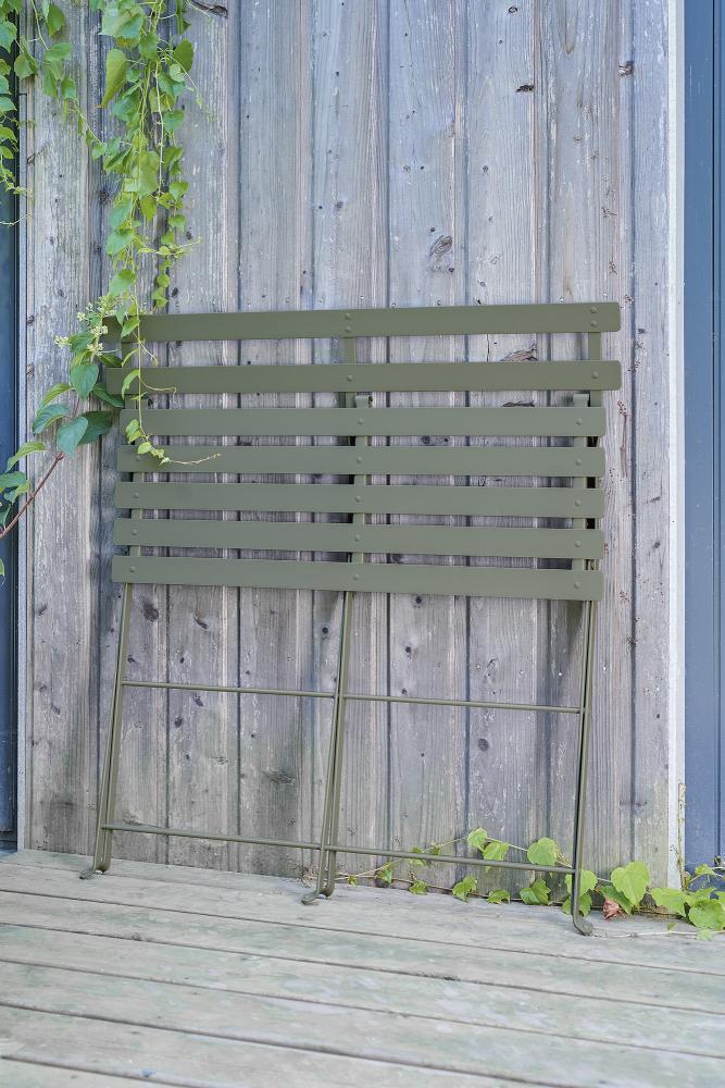 Olivegrüne Fermob Bistro Klappbank für zwei Personen, zusammengeklappt an einer Holzwand, Outdoor-Möbel für Garten und Balkon.