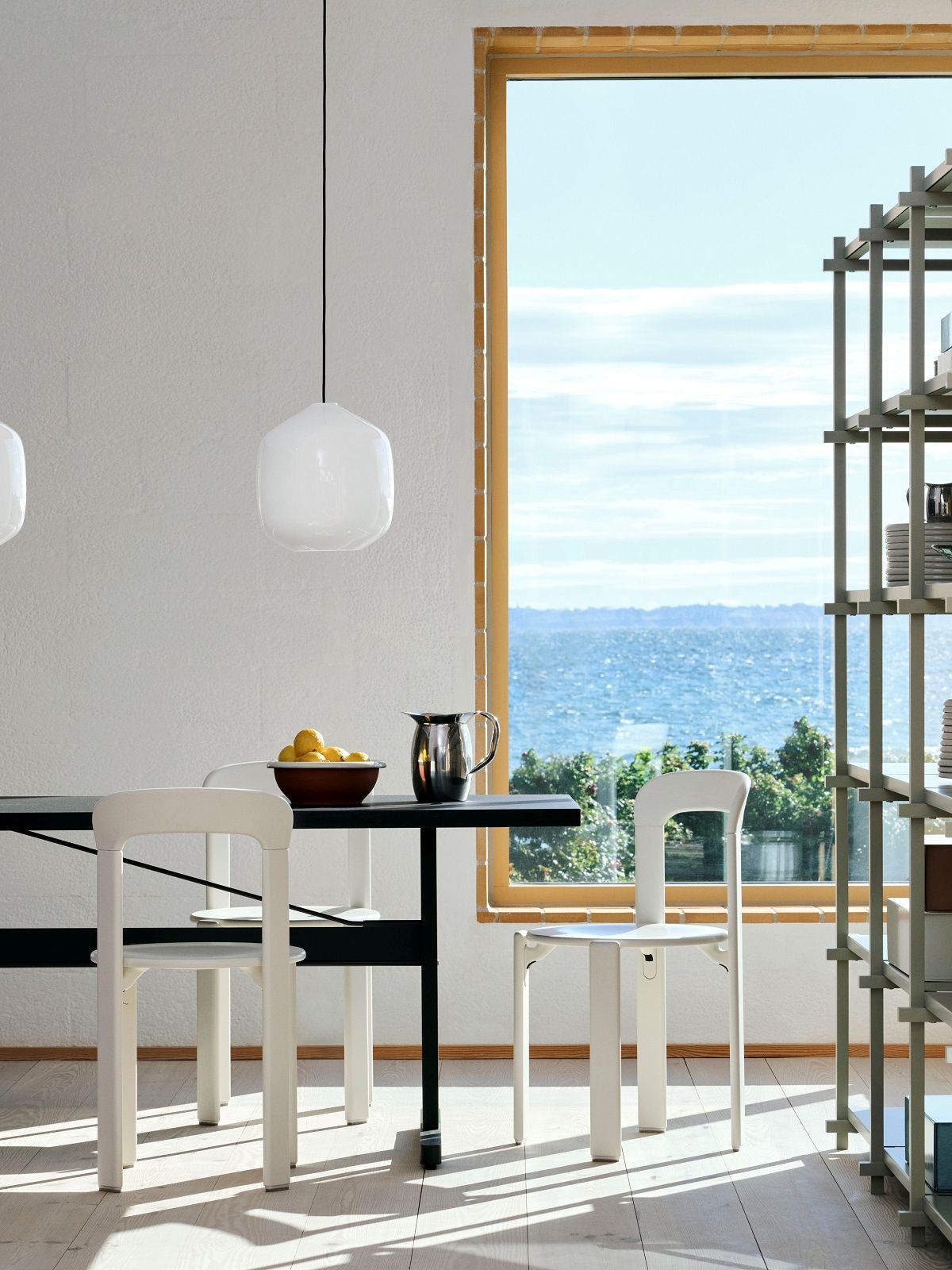 Helle Esszene mit Hay Buoy Pendelleuchte aus Glas, minimalistischem Tisch und Stühlen vor einem Fenster mit Meerblick.