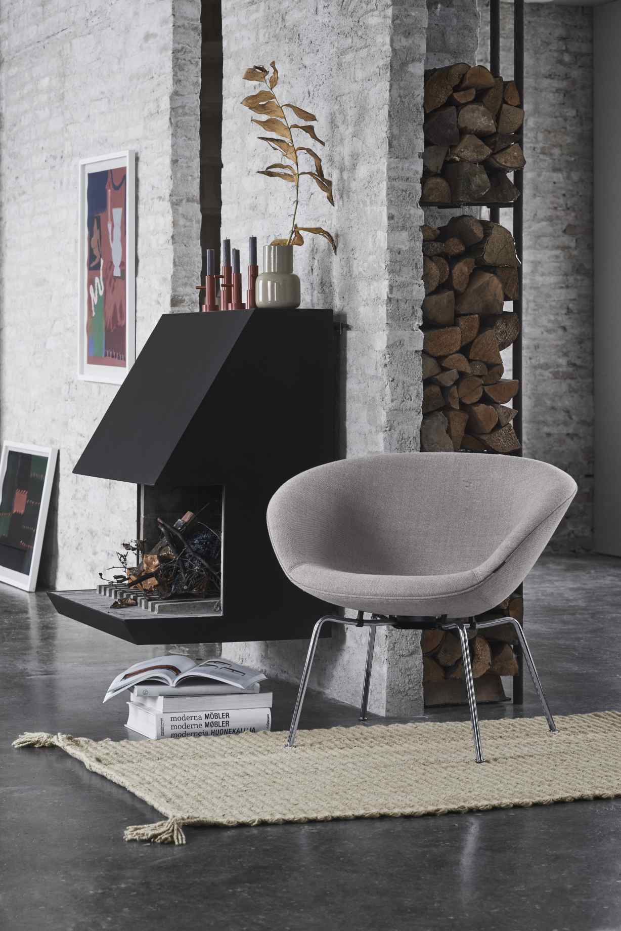 Grauer Fritz Hansen Pot Sessel mit Stahlbeinen vor Kamin und Brennholzregal in modernem Wohnzimmer.