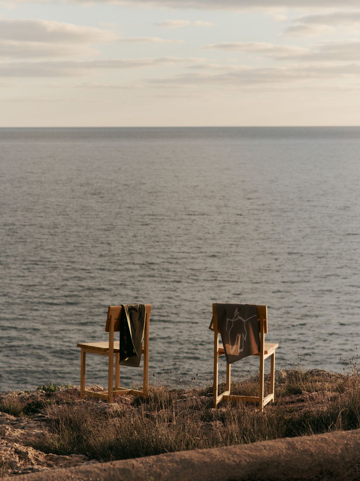 Zwei Ferm Living Esszimmerstühle aus Holz mit Handtüchern vor dem Meer, Außenbereich.